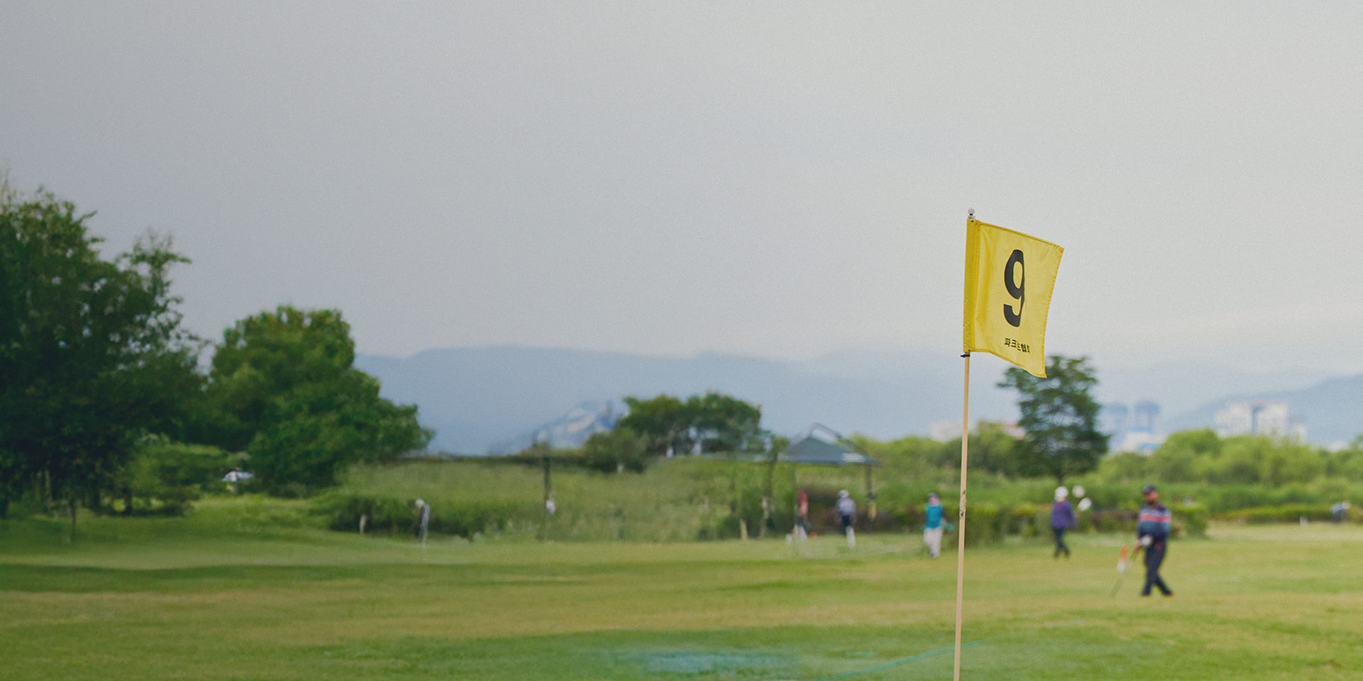 河川敷とみられる平坦なパークゴルフ場のグリーンに立つ9番のピンフラッグ。遠景に山と市街地