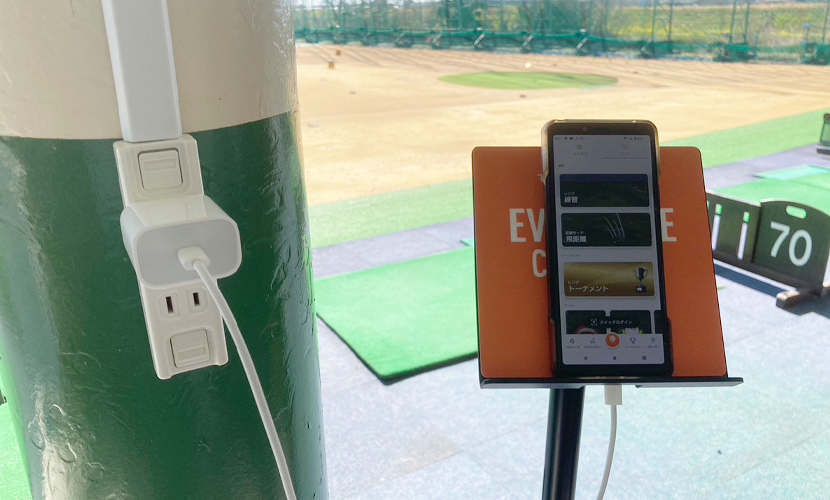リバーサイドゴルフ練習場 打席のスマホスタンドとコンセント、奥に天然芝のレンジ
